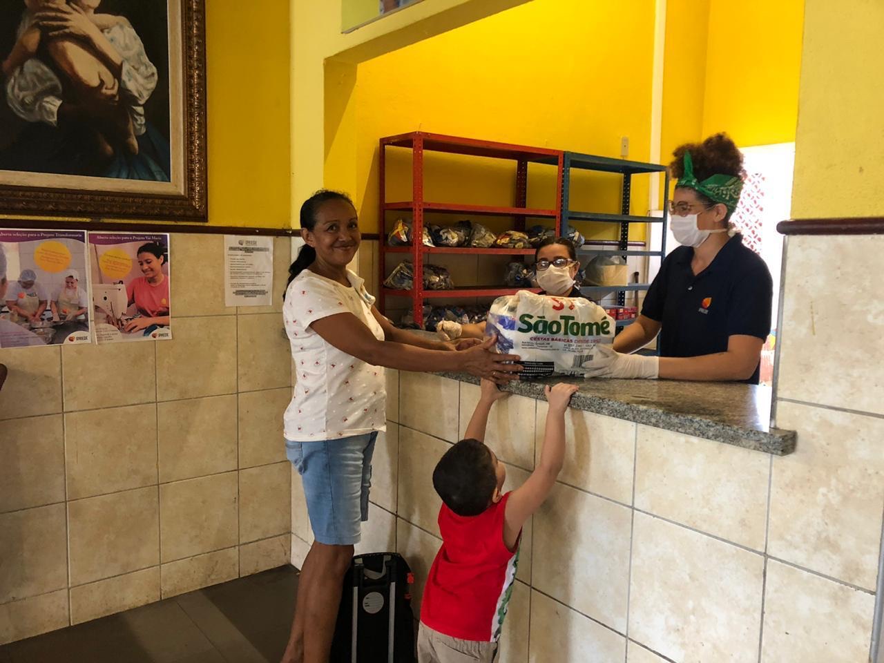 mulher de máscara em balcão entregando cesta básica para mulher sorrindo